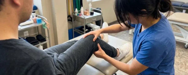 A physiotherapist examining a patient's knee joint stability, hands positioned on the knee, patient seated on a treatment bench in a bright modern clinic